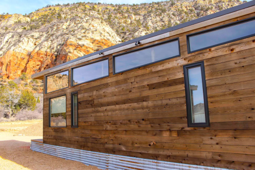 A modern wooden house with large windows, set against a backdrop of rocky cliffs and a clear blue sky.